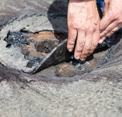 Worker lifts deteriorated roofing material during flat roof drainage system repair, exposing rusted metal and tar-coated layers. Closeup highlights inspection and maintenance of damaged roof component